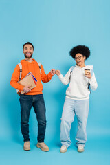 Canvas Print - full length of multiethnic students with notebooks and takeaway drink doing fist bump while smiling at camera on blue background.