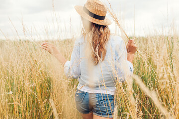 Wall Mural - Female fashion. Back view of young woman walking among high grasses in summer meadow wearing straw hat and linen shirt.