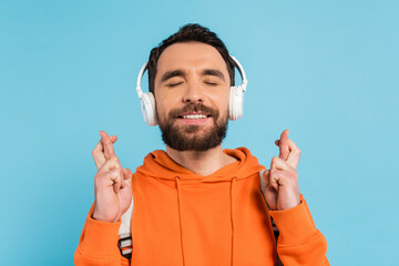 Wall Mural - positive student in wireless headphones holding crossed fingers while standing with closed eyes isolated on blue.