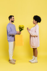 Wall Mural - full length of smiling bearded man gifting flowers to stylish african american woman on yellow background.