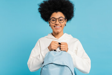 Wall Mural - happy african american student in white hoodie and eyeglasses holding backpack isolated on blue.