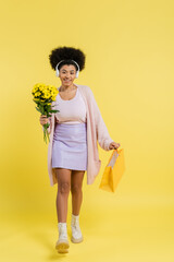 Wall Mural - full length of smiling african american woman in wireless headphones walking with flowers and shopping bag on yellow background.