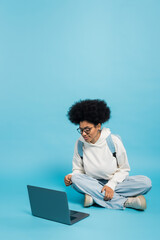 Wall Mural - full length of african american student in eyeglasses and white hoodie sitting with crossed legs near laptop on blue background.