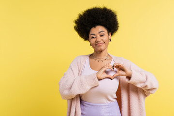 Wall Mural - positive african american woman in soft cardigan showing heart sign and looking at camera isolated on yellow.