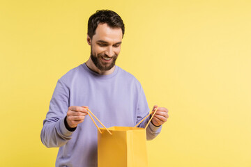 Wall Mural - pleased bearded man looking into shopping bag isolated on yellow.