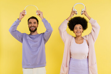Wall Mural - cheerful and trendy interracial couple holding wireless headphones above heads isolated on yellow.