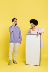 Wall Mural - african american woman pointing at phone template near smiling bearded man talking on mobile phone on yellow background.