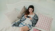Young woman using wireless technology lying on bed and chatting at home. Girl types on smartphone during breakfast.
