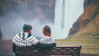 Back view of young beautiful couple sitting on bench near the Skogafoss waterfall in Iceland. Man and woman in lopapeysa