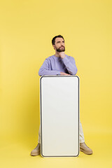 Wall Mural - thoughtful bearded man looking away near mock-up of white mobile phone on yellow background.