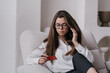 Anxious brunette European woman in glasses  holding credit card sitting in cozy chair, thinking about financial troubles, rent , debt. Upset caucasian girl loses her job. Money and financial crisis.