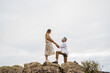 A young couple stands on a rocky cliff by the ocean, with the man down on one knee proposing to the woman