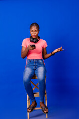 Wall Mural - Full length of cheerful young black woman with headphones and cellphone listening to music or audio book, sitting on chair