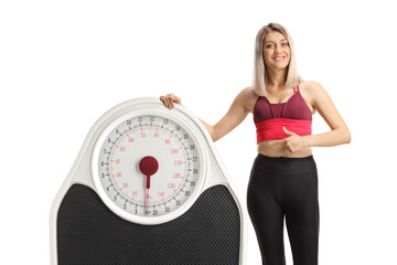 Poster - Young sporty woman leaning on a big weight scale and pointing