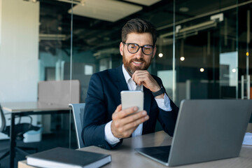 Business concept. Positive middle aged male entrepreneur using smartphone and modern laptop, sitting at desk in office