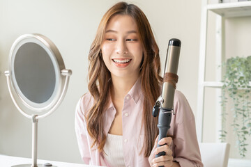 Beautiful curly brunette, attractive asian young woman, girl smiling using curler, curling iron long hair with ceramic curler straightener at home. Hairstyle and Hairdressing. Beauty and hair care.