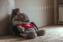 A Sad Nine-year-old Girl, She Sits In A Room In The Attic Without Repairs On The Floor, Hugged An Old Big Teddy Bear, Children's Anxieties And Sorrows