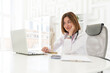 © Vitaliy - Portrait photo of young and beautiful smiling female doctor in white medical gown sitting and working on laptop in the office of the modern clinic. Online consultation and distant cure concept