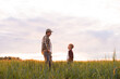 © Acronym - Farmer and his son in front of a sunset agricultural landscape. Man and a boy in a countryside field. Fatherhood, country life, farming and country lifestyle concept.