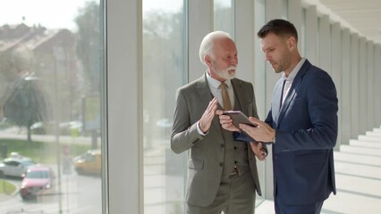 Wall Mural - Handsome mature businessman and his young colleague discussing financial report on digital tablet in the office corridor