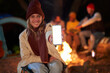 © CarlosBarquero - Young blonde girl showing a cell phone with a blank screen on the background of a campsite at night in outdoors. Smartphone in hand, millennial woman full of joy sitting in front of the bonfire.