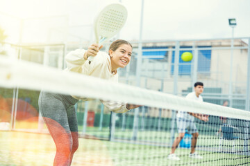 Wall Mural - Cheerful fit young girl paddle tennis player waiting to receive serve, ready to perform forehand to return ball to opponent field on outdoor court on summer day