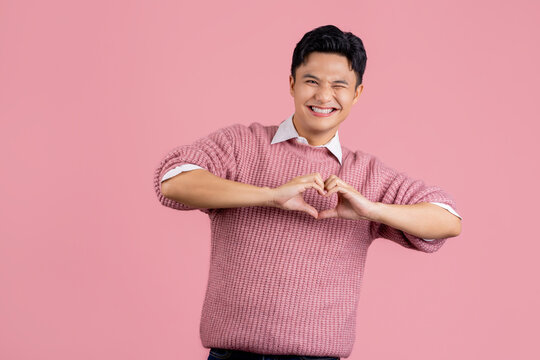 Young asian man wearing casual pink sweater smiling in love doing heart symbol shape with hands. romantic concept.