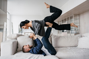 Happy Italian beardy man in casual laying on couch lifting up girlfriend by legs celebrating new apartment. Excited young hispanic couple having fun at hotel room on vacations. Newlyweds at honeymoon.