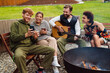 © Drobot Dean - Cheery friends drinking tea and using smartphone while resting around campfire with guitar