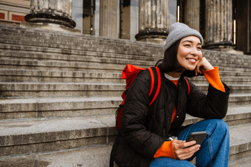 Poster - Positive asian woman tourist using mobile phone while sitting on old museum stairs