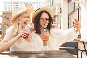 Wall Mural - Two young beautiful smiling hipster female in trendy summer straw hats. Sexy carefree women holding and drinking fresh vegetable cocktail smoothie drink in plastic cup with straw. Taking selfie