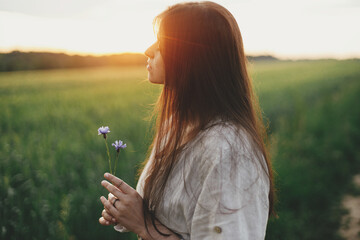 Poster - Woman relaxing in wheat field in warm sunset light. Tranquil atmospheric moment. Portrait of young female in rustic dress holding wildflowers in hands in evening summer countryside