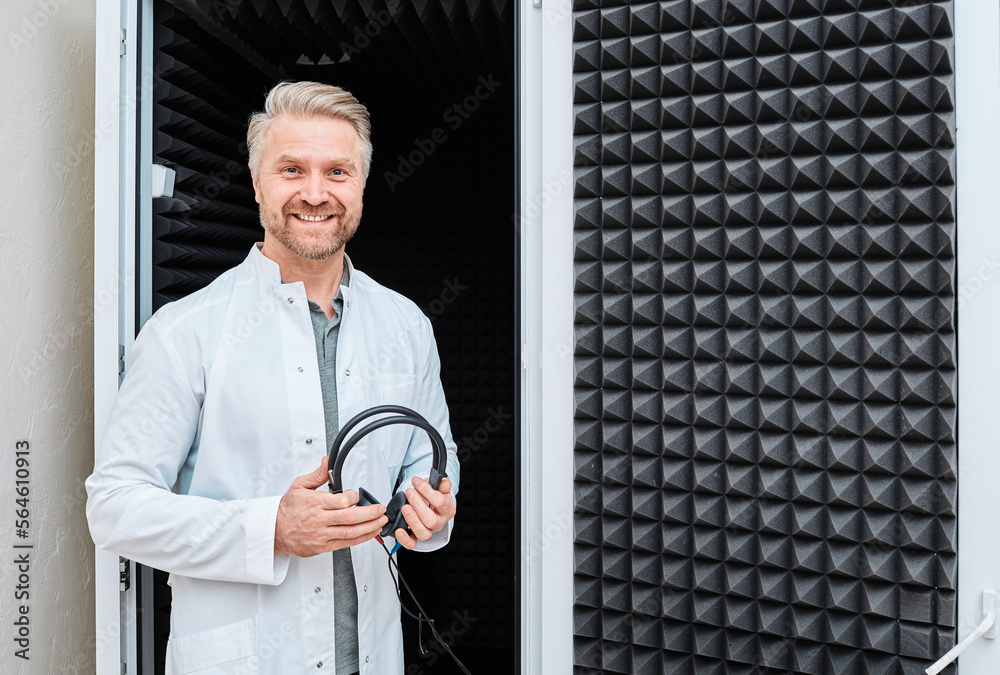 Hearing specialist standing near soundproof audiometric booth with audiometer headset in hands during audiometry exam at hearing center