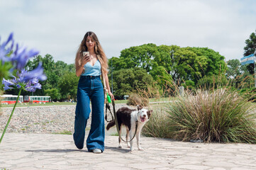 young latin woman using the phone and walking with her dog in the park