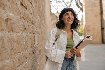 Wall Mural - Image of happy student in hands notebooks staying near to wall with backpack. Caucasian brunette woman wearing casual clothes. Concept of lifestyle