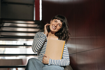 Wall Mural - Smiling pretty cute woman with workbooks in working place look at camera. Wearing striped shirt, blue jeans touching hair. Lifestyle concept 