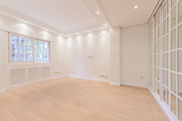 Empty bedroom with white painted walls, light wooden floor, French-style aluminum windows, radiator covers of the same material and wardrobes with aluminum sliding doors and mirrors