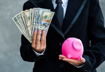 business woman in black suit keeping money dollar banknotes in hand put in pink piggy bank. Insert banknotes dollar in piggy bank saving box.