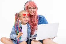 Adorable Daughter With Funny Colorful Braids Wearing Denim Clothes Watching Cartoons On Laptop. Young Mother Wearing Pink Wig And Headphones Sitting On White Background In Studio Copyspace.