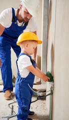 Man and little boy installing cable canals and sockets in wall. Male electrician and kid in safety construction helmet and work overalls mounting electrical wiring in apartment under renovation.