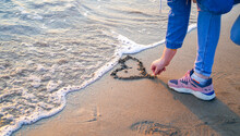 Woman Drawing Heart On The Beach Sand Near Mediterranean Sea's Foamy Waves
