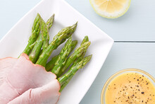 Fresh Cooked Green Asparagus With Ham Slices, Hollandaise Sauce On The Side, Photographed Overhead On Light Blue Wood (Selective Focus, Focus On The Top Asparagus Heads, The Ham And The Sauce)