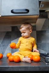 A little boy 1 year old in yellow clothes is sitting in the kitchen on the table with a plate of orange tangerines. Portrait of a cute one-year-old boy and sweet citrus fruits.