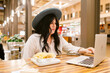 © ByLorena/Stocksy - Woman eating noddle while using laptop in food court