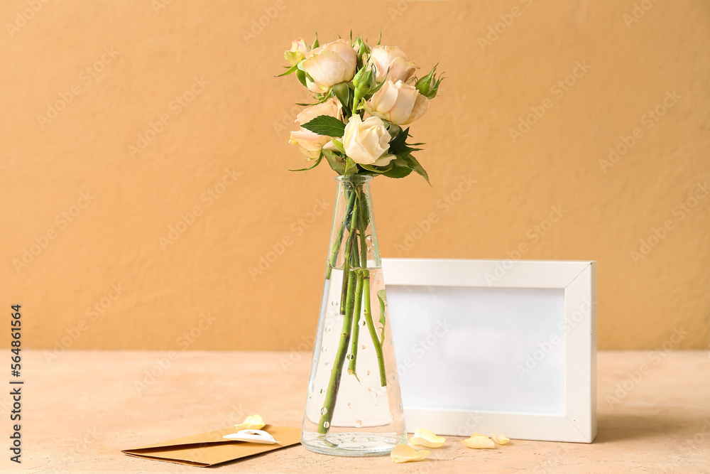 Vase with beautiful rose flowers, envelope and empty picture frame on table near color wall