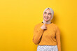 Beautiful thoughtful Asian girl wearing hijab looking into empty space, touching chin, thinking about something interesting isolated on yellow background