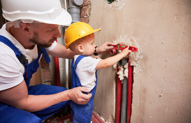 Man and little boy using spirit level tool while installing plumbing pipes in apartment. Male plumber and child in safety construction helmet measuring pipes level with building instrument.