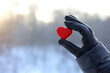 Red knitted heart in female hand in black leather glove on snow park and sun background. Concept of romantic love, Valentine's day, winter weather