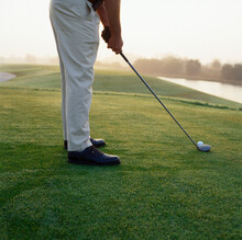 Man Stands With Golf Club And Ball On Grass Preparing To Tee Of In Early Morning Haze.