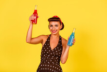 Smiling Lady In Polka Dot Dress And Retro Hairstyle Holds Bottles Of Soda Drink In Her Hands On Yellow Background. A Pin-up Look With A Modern Twist. Vintage Party